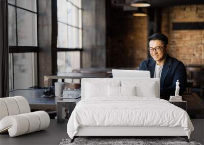 Asian businessman typing on laptop during work in cafe. Concept of remote and freelance work. Smiling adult successful man wearing suit and glasses sitting at wooden desk. Sunny day Wall mural