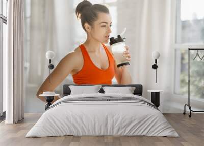 Athletic woman in bright sportswear drinking protein beverage from a shaker, standing indoors. The concept of sports nutrition during exercise Wall mural