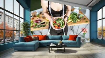 Athletic woman in sportswear holding salad near the table full of healthy products. Healthy eating and sports lifestyle concept Wall mural