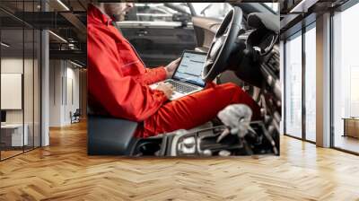 Auto mechanic in red uniform diagnosing car with computer sitting on the driver seat at the car service Wall mural