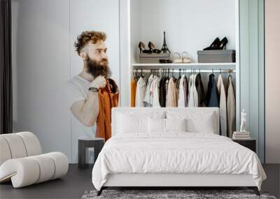 Bearded man trying shirt, choosing clothes to wear in the wardrobe at home Wall mural