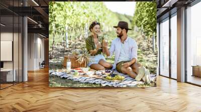 Beautiful couple having romantic breakfast with lots of tasty food and wine, sitting together on the picnic blanket at the vineyard on a sunny morning Wall mural