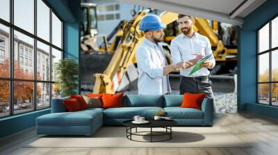 Builder choosing heavy machinery for construction with a sales consultant standing with some documents on the open ground of a shop with special vehicles Wall mural