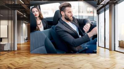 Businesswoman on the backseat with elegant man driving a car in the city. Worried about a delay to an important meeting Wall mural