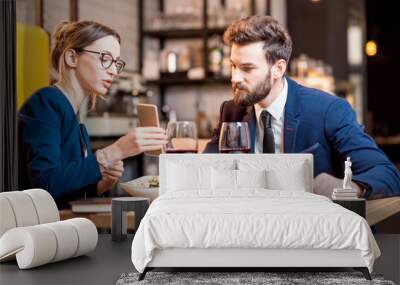 Caucasian business couple dressed strictly in the suits working together with smart phone sitting at the restaurant during the dinner Wall mural