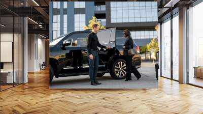 Chauffeur helps business couple to get in the car, opening a door of a luxury black taxi. Man and woman having a business trip together Wall mural