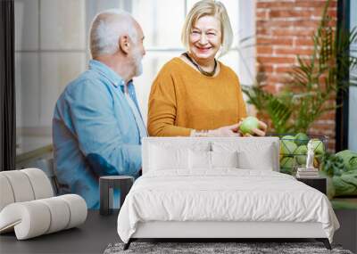 Cheerful senior couple eating fruits standing together with healthy food on the kitchen at home. Concept of healthy nutrition in older age Wall mural