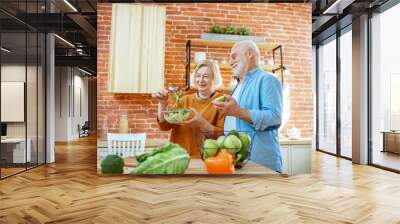 Cheerful senior couple eating salad standing together with healthy food on the kitchen at home. Concept of healthy nutrition in older age Wall mural