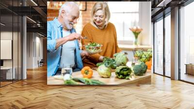 Cheerful senior couple eating salad standing together with healthy food on the kitchen at home. Concept of healthy nutrition in older age Wall mural