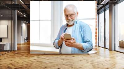 Cheerful senior grandfather using phone standing near the window at home Wall mural