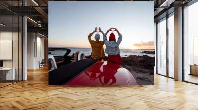 Couple enjoying beautiful views on the ocean, standing together near the car on the rocky coast, showing with hands heart shape. Carefree lifestyle, love and travel concept Wall mural