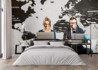 Couple of travel managers working online with laptops and headsets at the agency office with world map on the background Wall mural