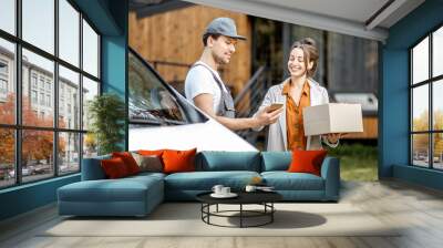 Delivery company employees in uniform delivering goods to a client by car, woman signing on a smartphone while receiving parcel near home Wall mural