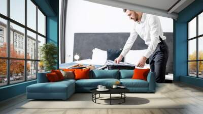 Elegant businessman packing his clothes into the suitcase on the bed of the hotel room during the business trip Wall mural