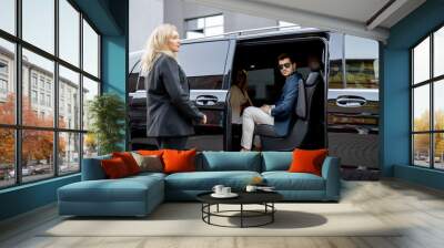 Female chauffeur helps a business man to get out of vehicle, opening a door of luxury black minivan. Concept of personal driver, luxury taxi for business delegation Wall mural