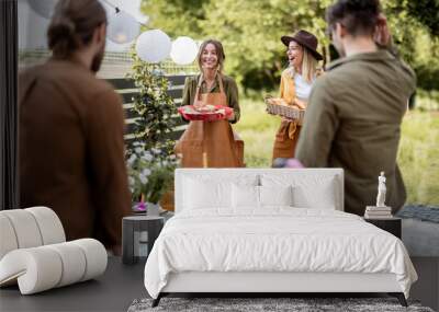 Friends preparing for the festive dinner, two housewifes carrying hot meal and serving the table. A group of young people gathering at backyard of the house in nature Wall mural