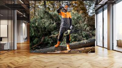 Full length portrait of a professional lumberjack in protective workwear standing with a chainsaw on the logs in the forest Wall mural