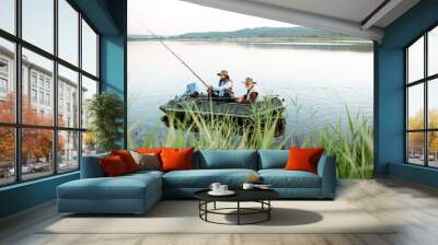 Grandfather with adult son fishing on the inflatable boat on the lake with green cane on the foreground early in the morning Wall mural