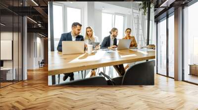 Group of a young office employees dressed casually in the suits having some office work at the large meeting table in the bright sunny room Wall mural