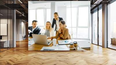 Group of a young office employees dressed casually in the suits having some office work at the large meeting table in the bright sunny room Wall mural