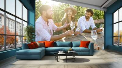 Group of a young people drinking wine and having fun together while sitting at the dining table outdoors on the vineyard on a sunny evening Wall mural