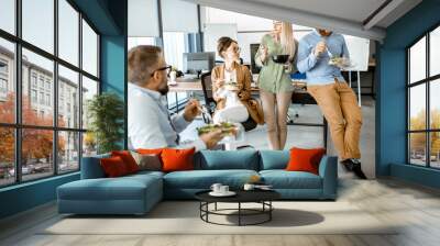 Group of diverse colleagues eating takeaway salad, sitting together and having fun during a lunchtime in the office Wall mural