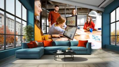 Group of young coworkers dressed casually working together on the computers with some charts sitting in the modern office interior Wall mural