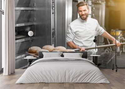 Handsome baker in uniform taking out with shovel freshly baked buckweat bread from the oven at the manufacturing Wall mural