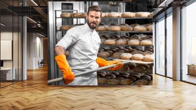 Handsome baker in uniform with orange working gloves putting with shovel from the oven bread loafs on the shelves at the manufacturing Wall mural