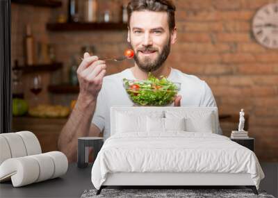 Handsome bearded man in white t-shirt eating salad with tomatoes in the kitchen. Healthy and vegan food concept Wall mural