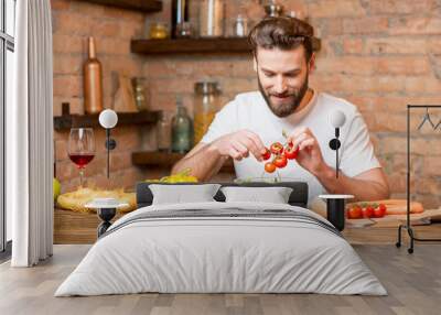 Handsome bearded man in white t-shirt making salad with tomatoes and pepper in the kitchen. Healthy and vegan food concept Wall mural