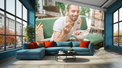 Handsome man eating healthy salad sitting indoors on the green sofa with bag full of vegetables on the background. Healthy eating concept Wall mural