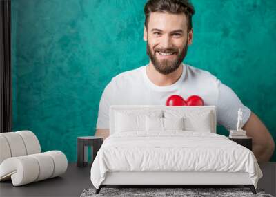 Handsome man in the white t-shirt holding red heart on the painted green wall background. Valentine's Day concept Wall mural