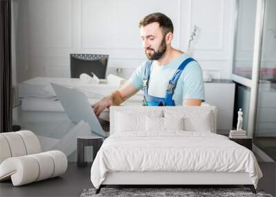 Handsome repairman or foreman in uniform working with laptop and architectural drawings at the office Wall mural