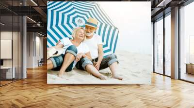 Happy senior couple relaxing, lying together under umbrella on the sandy beach, enjoying their retirement near the sea Wall mural