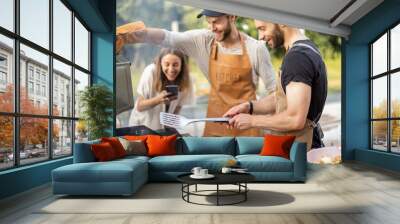 Happy young friends hanging out together, grilling meat and buns for burgers on a modern grill at picnic. People cooking food outdoors Wall mural