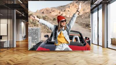 Lifestyle portrait of a carefree woman dressed casually in jeans and red hat sitting on the car trunk, enjoying road trip on the desert valley Wall mural