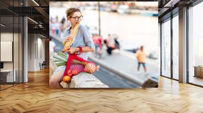 lifestyle portrait of a young woman with mesh bag full of fresh food eating baguette oudoors in the  Wall mural