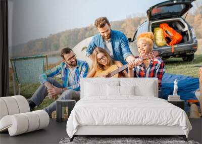 Multi ethnic group of friends dressed casually having fun playing guitar during the outdoor recreation with tent, car and hiking equipment near the lake Wall mural