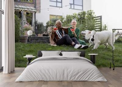 Multiracial couple eating watermelon in their garden. Concept of relationship and enjoying time together with a dog. Black man hugging his european girlfriend. People covered in plaid Wall mural