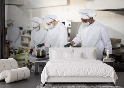 Multiracial team of cooks in uniform and face masks cooking meals for a restaurant in the kitchen. Concept of teamwork at restaurant during pandemic. Latin, Asian and European guys cooking together Wall mural