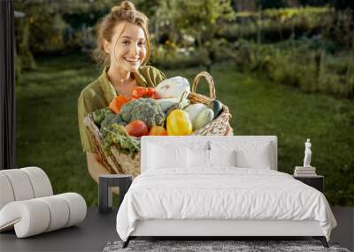 Portrait of a beautiful young woman with basket full of freshly picked up vegetables standing in the garden on a sunny evening Wall mural