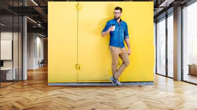 Portrait of a handsome man in blue t-shirt standing with coffee to go on the yellow background Wall mural