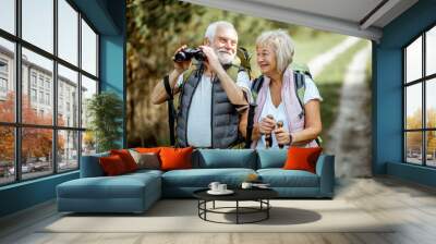 Portrait of a happy senior couple standing together with binoculars, backpacks and trekking sticks while hiking in the forest. Concept of an active lifestyle on retirement Wall mural