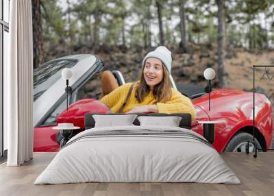 Portrait of a stylish woman in yellow sweater sitting on the driver's seat of convertible sports car on the roadside. Carefree lifestyle and traveling concept Wall mural