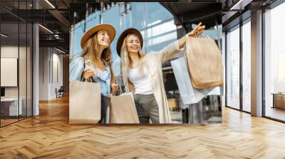 Portrait of a two happy women with shopping bags, pointing forward while standing together in front of the shopping mall Wall mural