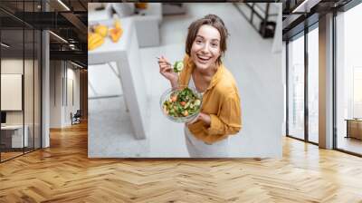 Portrait of a young and cheerful woman dressed in bright shirt eating salad at home. Concept of wellbeing, healthy food and homeliness Wall mural