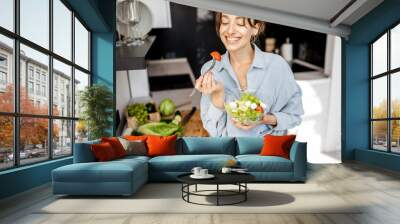 Portrait of a young and cheerful woman eating healthy salad on the kitchen at home. Healthy eating, food and lifestyle concept Wall mural