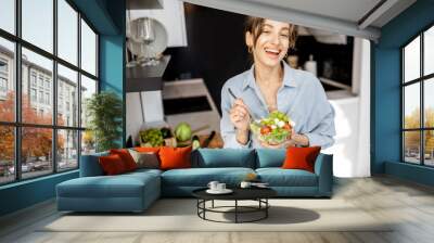 Portrait of a young and cheerful woman eating healthy salad on the kitchen at home. Healthy eating, food and lifestyle concept Wall mural