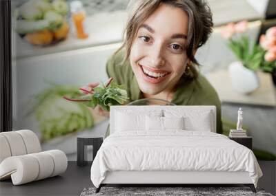 Portrait of a young and cheerful woman eating salad standing on the kitchen with food ingredients on the background, view from above. Vegetarianism, wellbeing and healthy lifestyle concept Wall mural